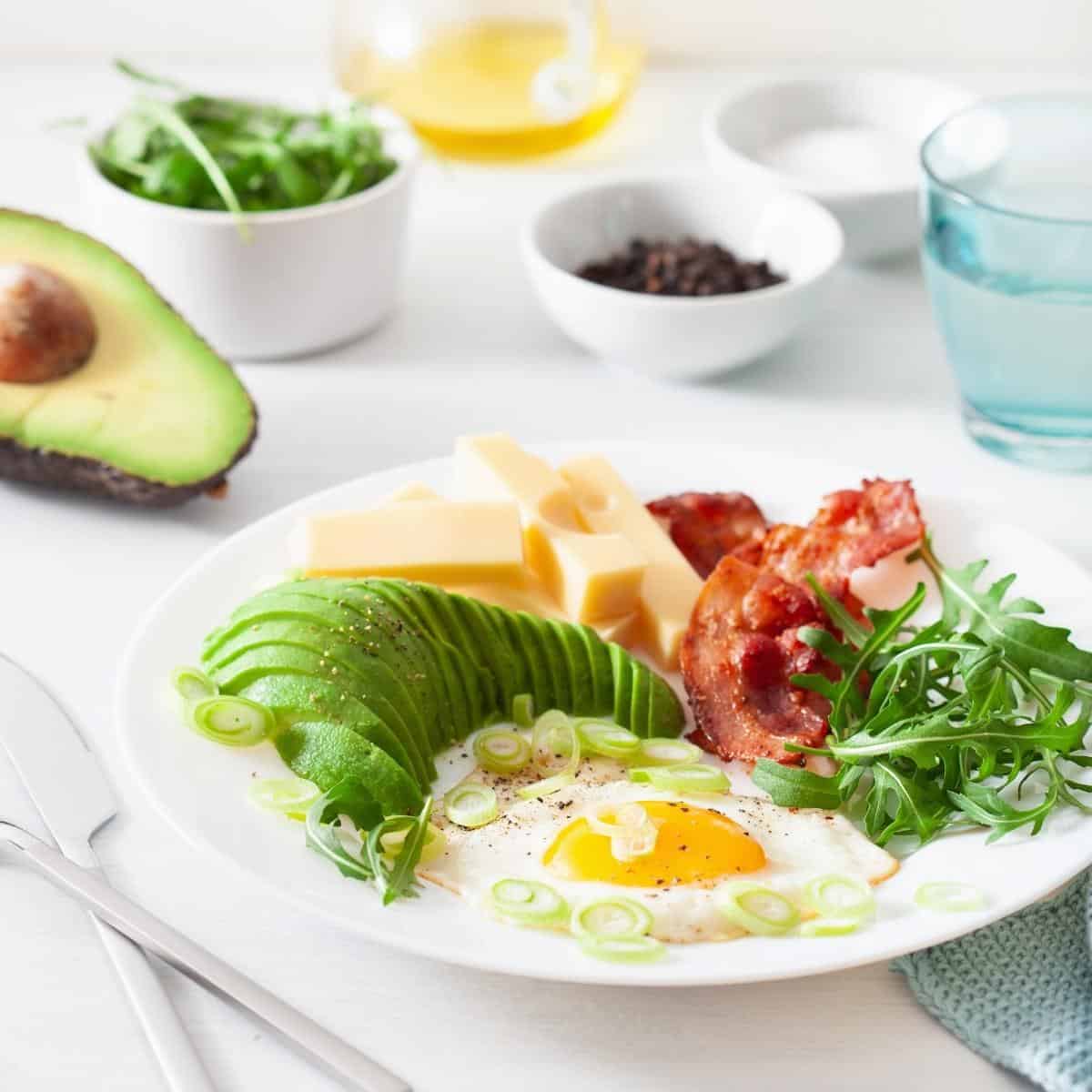 A white plate with eggs, bacon, cheese, avocado, arugula and other keto breakfast items.