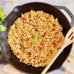 A skillet with sautéed corn and bacon garnished with a sprig of cilantro, accompanied by a wooden spoon.