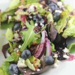 A fresh mixed greens salad with blueberries, feta cheese, and red onions on a white plate.