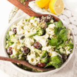 A bowl of broccoli salad with grapes, seeds, and a creamy dressing, garnished with parsley, served with lemon wedges on the side.