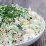 A bowl of creamy macaroni salad garnished with chopped herbs on a wooden surface.