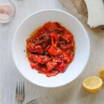 A bowl of roasted red peppers seasoned with herbs, accompanied by slices of cheese, lemon halves, and spices on a wooden kitchen table.