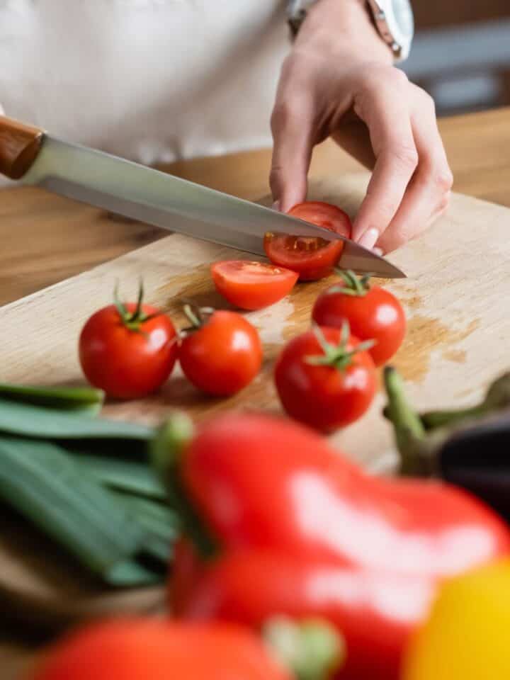 A person slicing tomatoes on a wooden cutting board with various vegetables nearby for mise en place.