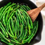 Sautéed green beans in a cast iron skillet with a wooden spatula.