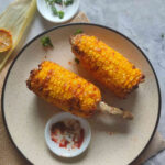 Two grilled corn cobs on a plate with a sprinkle of spices and a wedge of lime on the side.