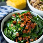 A bowl of sautéed greens topped with bacon bits, with cornbread and a dish of beans in the background.