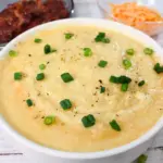 A bowl of creamy soup garnished with green onions and black pepper, with shredded carrots and a dish with a brown food item in the background.