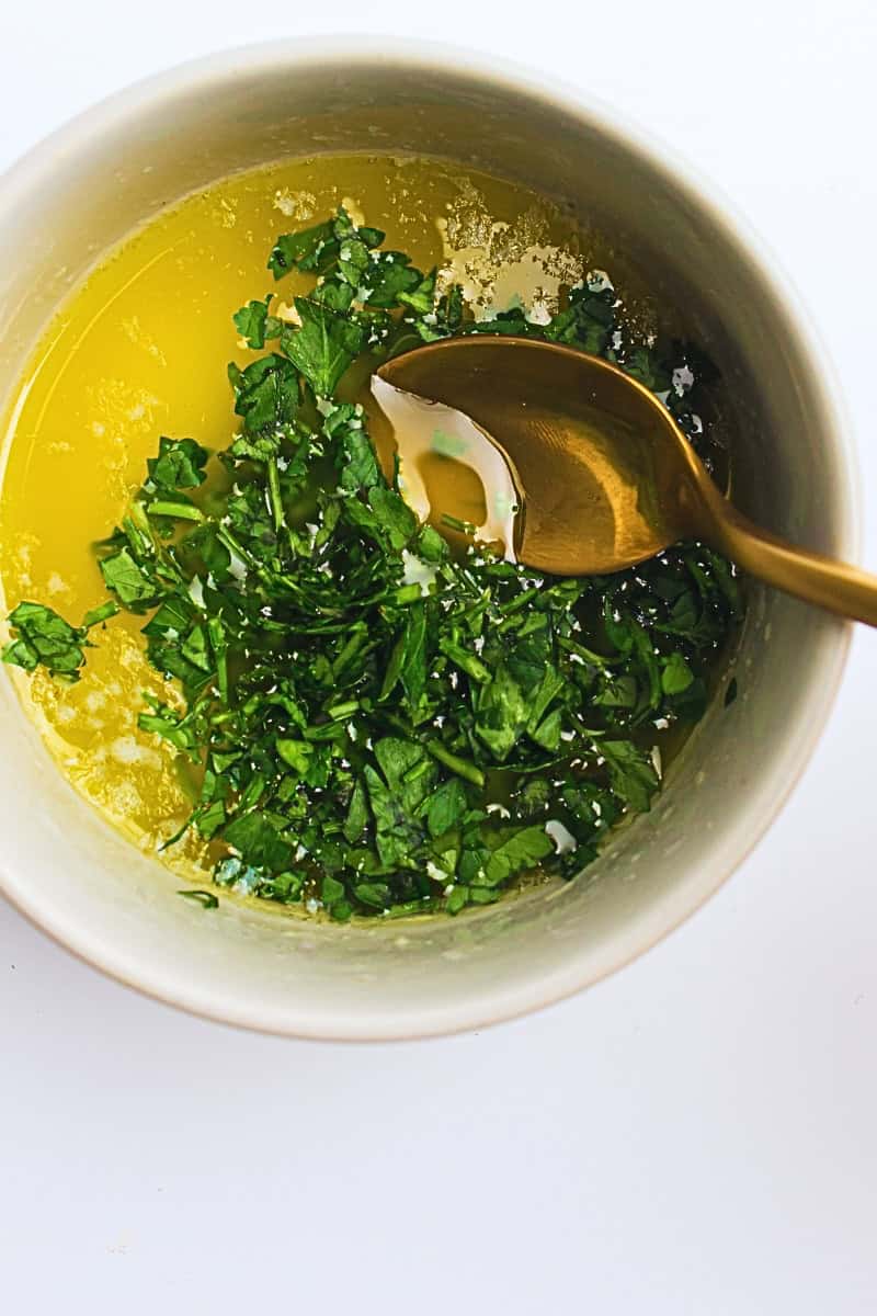 A bowl containing oil with chopped herbs and a spoon, set against a white background, perfect for seasoning air fryer steak bites.