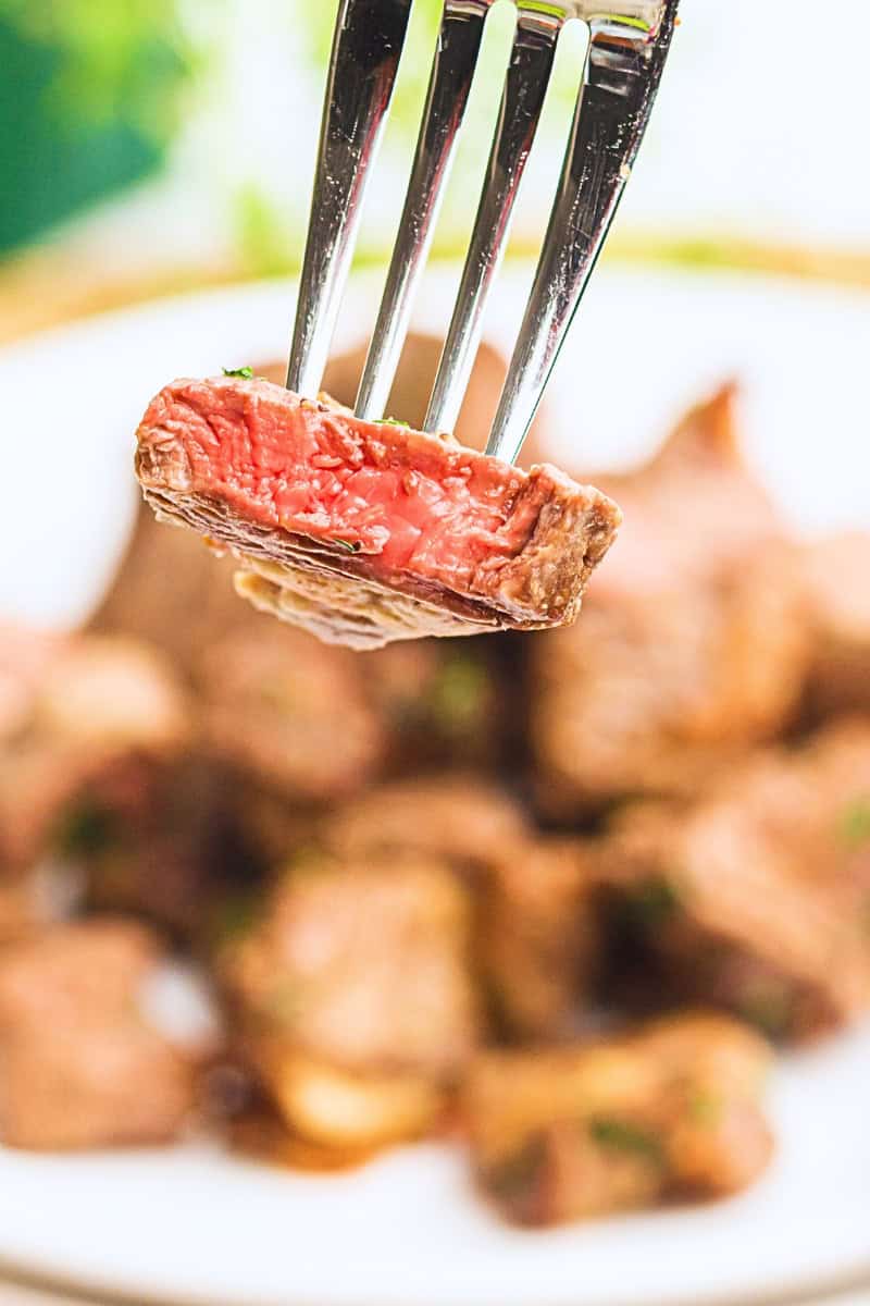 A piece of medium-rare air fryer steak bites held by a fork in focus with more steak pieces blurred in the background on a plate.