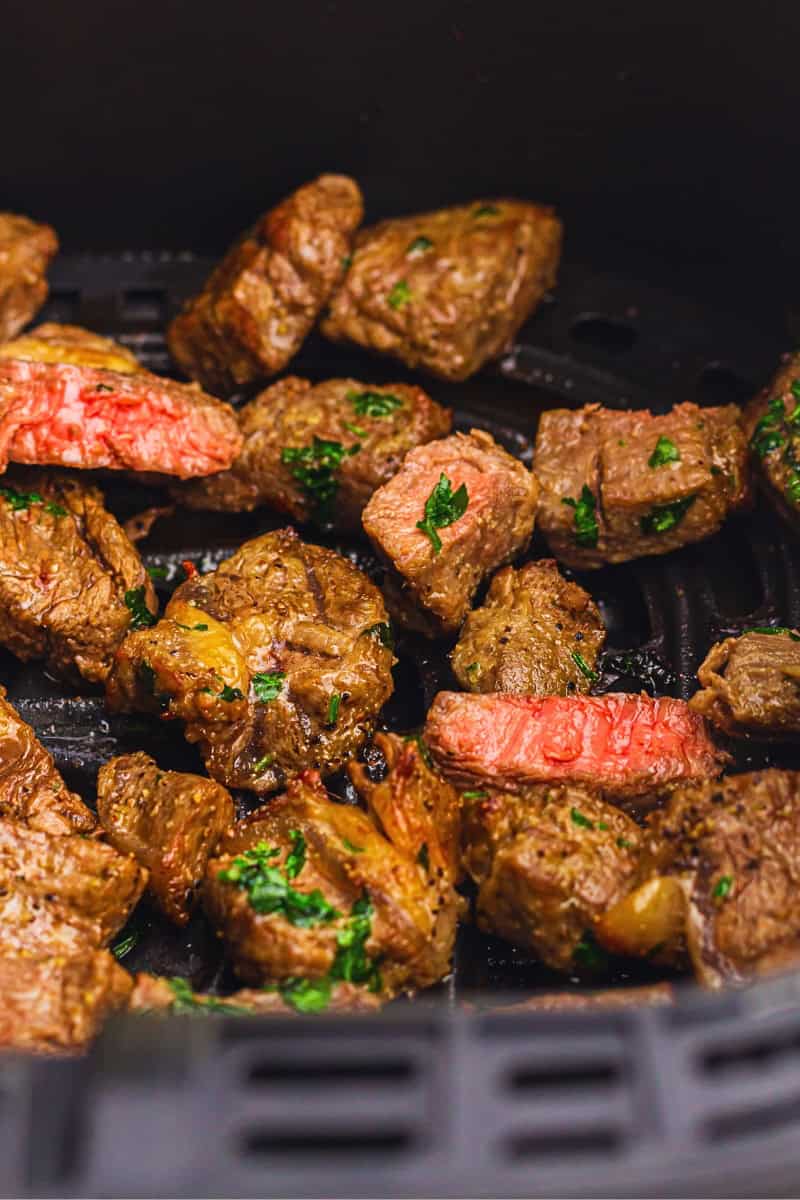 Grilled beef chunks with herbs in an air fryer basket, showcasing varying degrees of doneness.