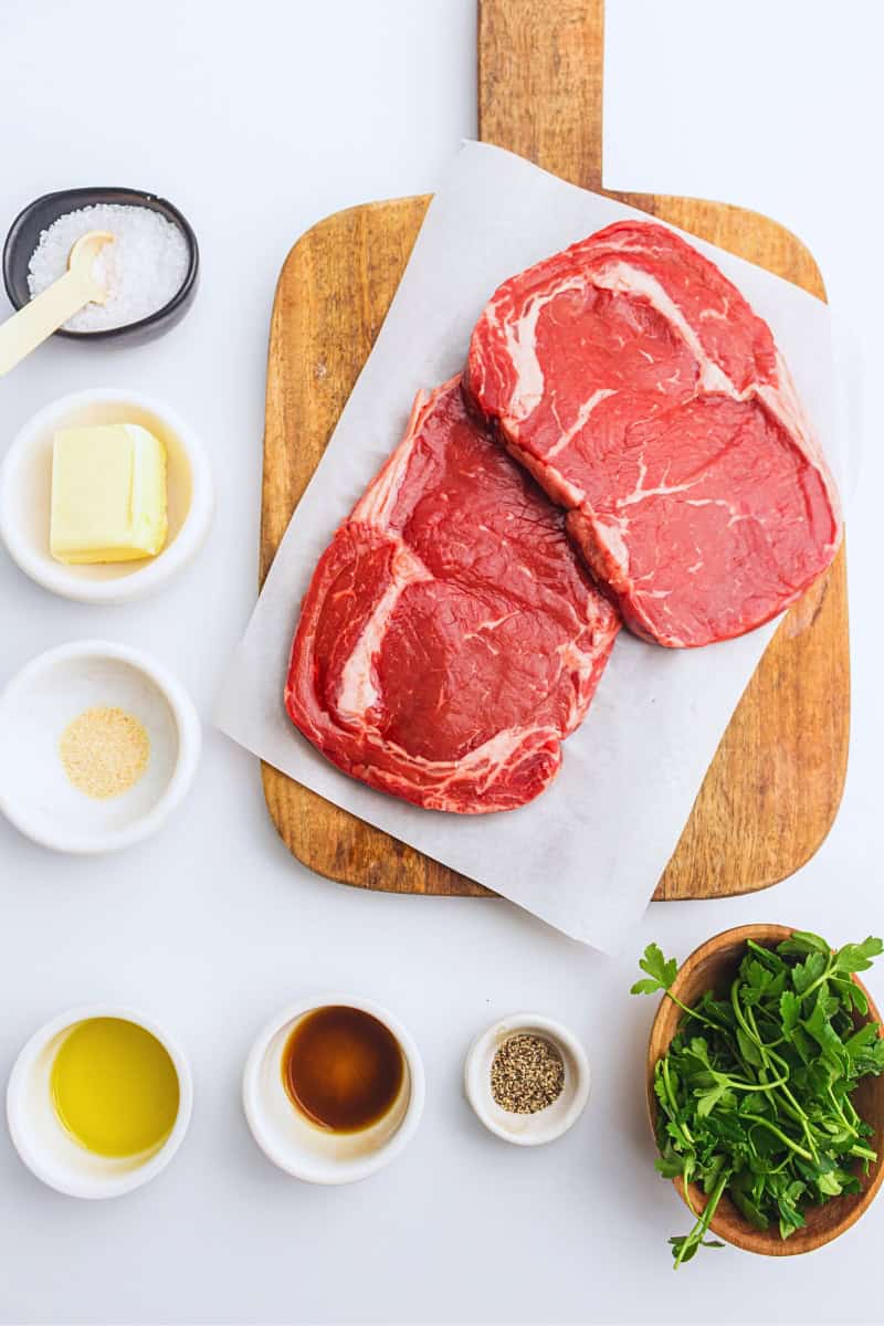 Two raw ribeye steaks on a wooden cutting board, surrounded by small bowls with salt, pepper, oil, butter, and arugula on a white background, ready for cooking as air fry